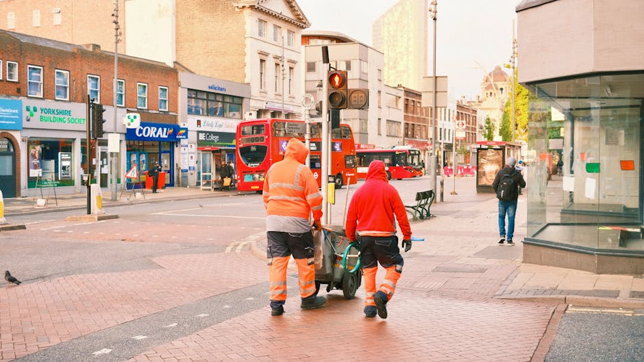 Two professional cleaners from Cleaners Watford are performing surface cleaning in an urban area, pushing a cleaning cart along the paved sidewalk. One cleaner is dressed in a grey and orange uniform,